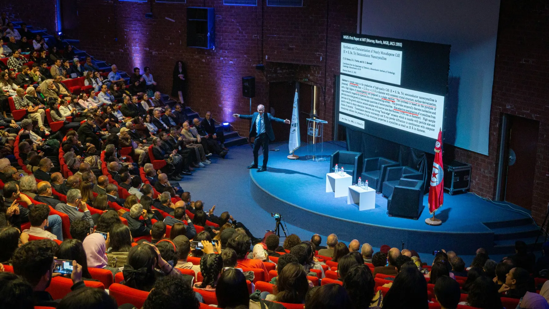 A speaker presenting beside a large screen in a full university lecture theatre.